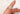 A close-up of a hand with light purple nail polish wearing a gold three-stone engagement ring, featuring a large emerald-cut center diamond flanked by two smaller trapezoid-cut diamonds, set against a soft white background.